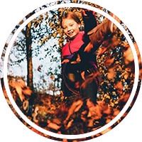 Kind springt lachend durch fallende Herbstblätter während eines Familien-Outdoorshootings im Wald.